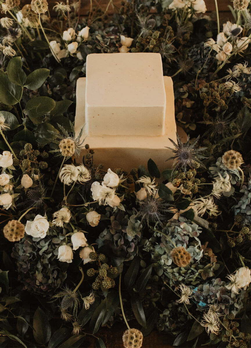 A two-tier, square wedding cake covered in buttercream set in a field of roses, hydrangea, thistle, and scabiosa pods.