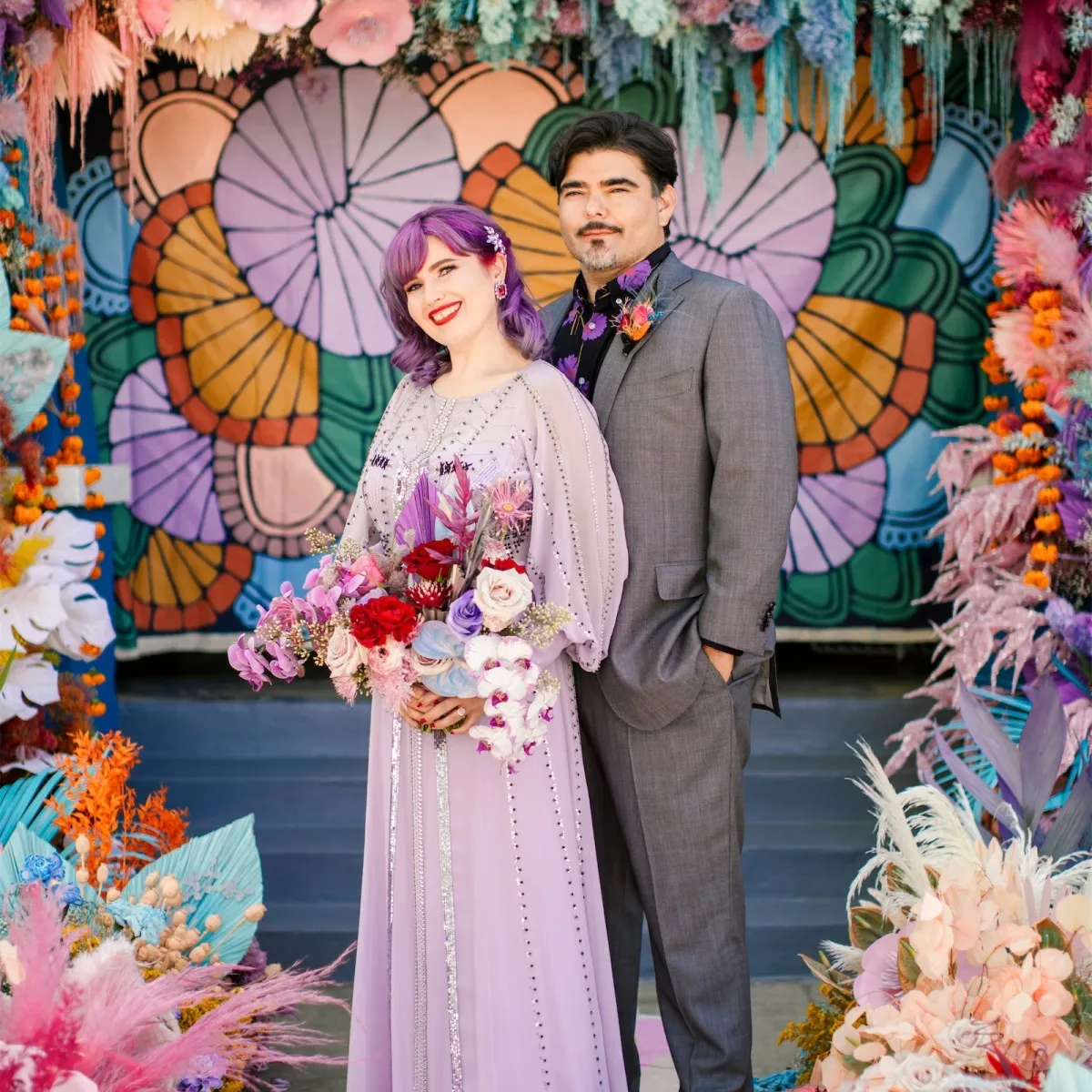 A bride and groom pause for a portrait at their vibrant outdoor wedding, which incorporated a rainbow floral installation and painted backdrop at their ceremony.