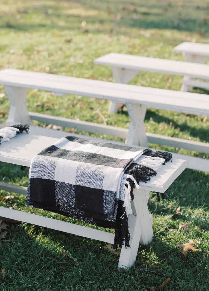 Wedding Aisle: A black and white gingham blanket folded on a white bench at an outdoor wedding ceremony.