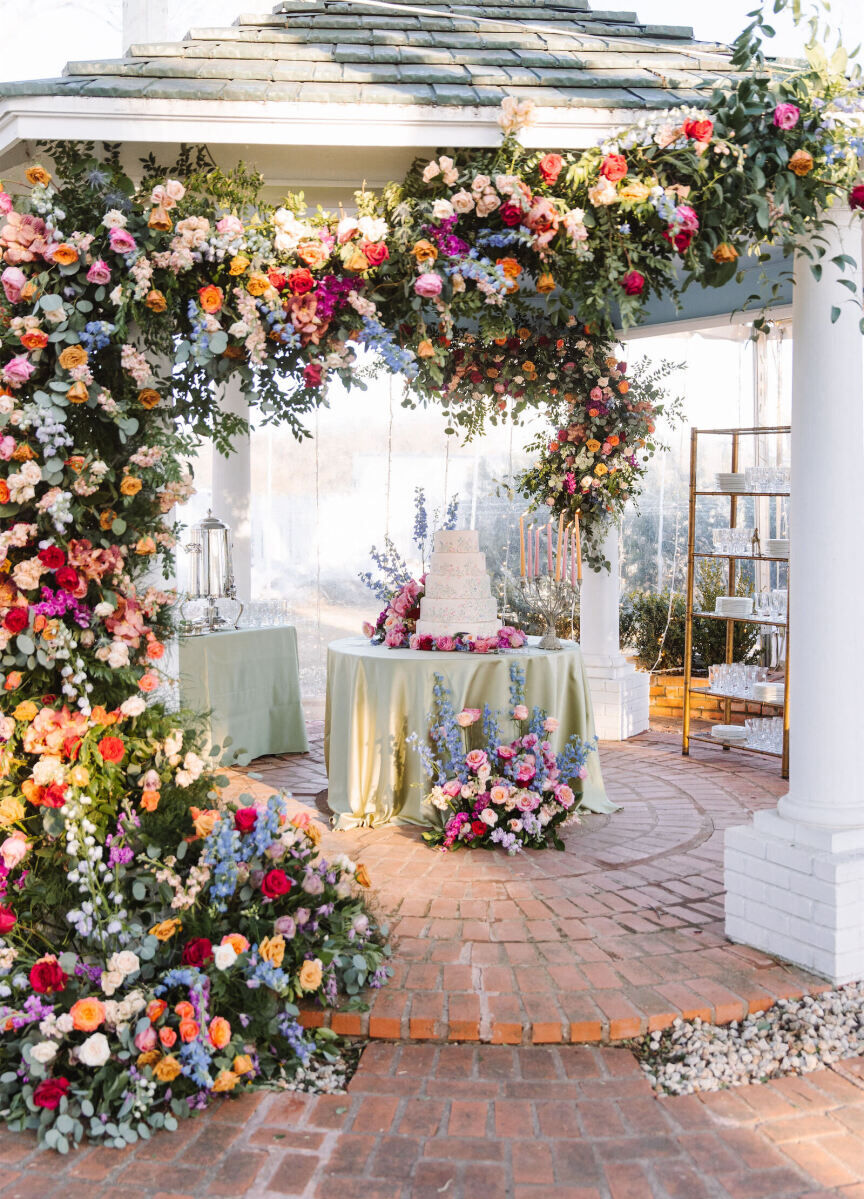 Wedding cake on display table under gazebo decorated with colorful florals.