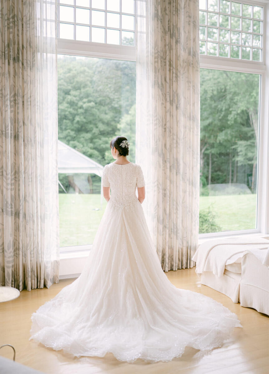 Wedding details: Bride staring out a window, wearing a classic white weddings dress featuring a cascading train