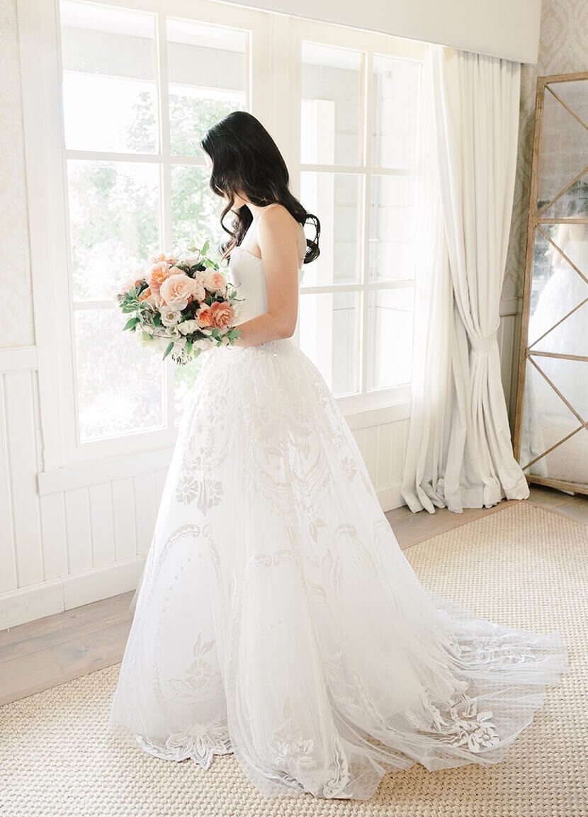 Wedding Dress Shopping: A bride looking down and holding a pink floral bouquet while wearing a strapless gown indoors.