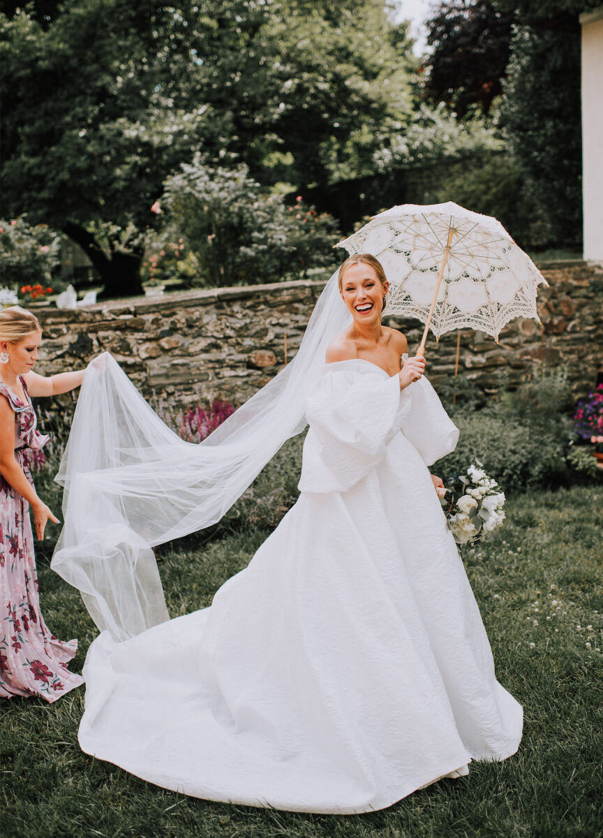 Wedding Dress Shopping: A bride smiling with a parasol as her bridesmaid adjusts her floor-length veil.