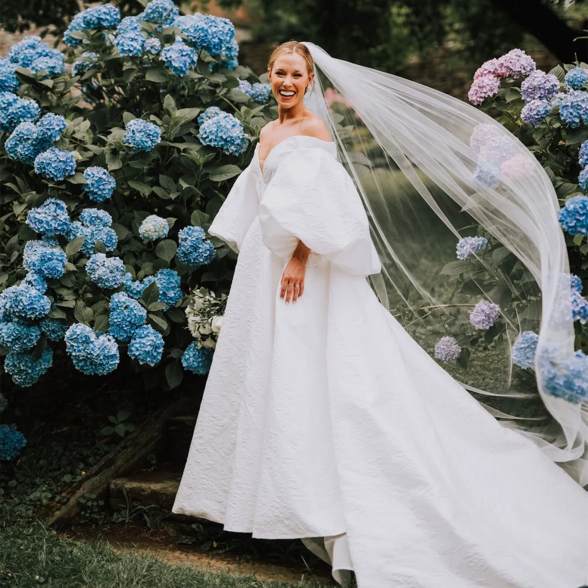 Wedding Dress Shopping: A bride smiling near blue hydrangeas, wearing an off-the-shoulder gown and floor-length veil.