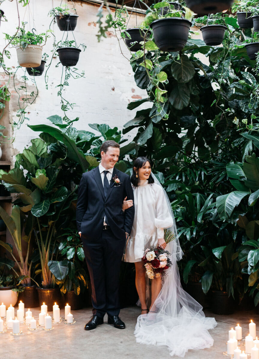 Wedding Dress Shopping: A bride holding a multi-colored bouquet with her arm linked to the groom, and both of them are in an outdoor patio with candles on the ground.