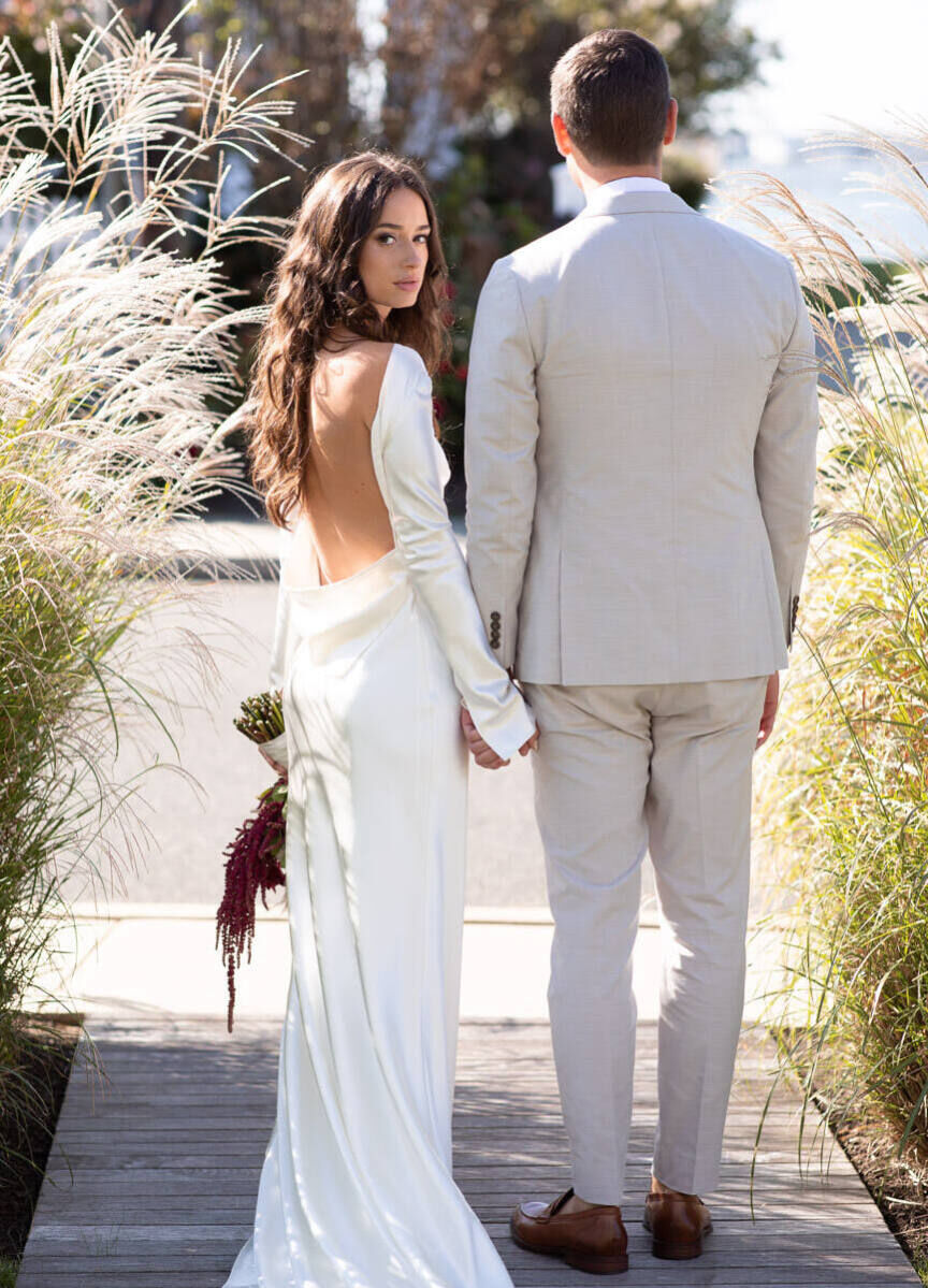 Wedding Dress Shopping: A bride looking over her shoulder in a low-back wedding gown while holding hands with her groom.