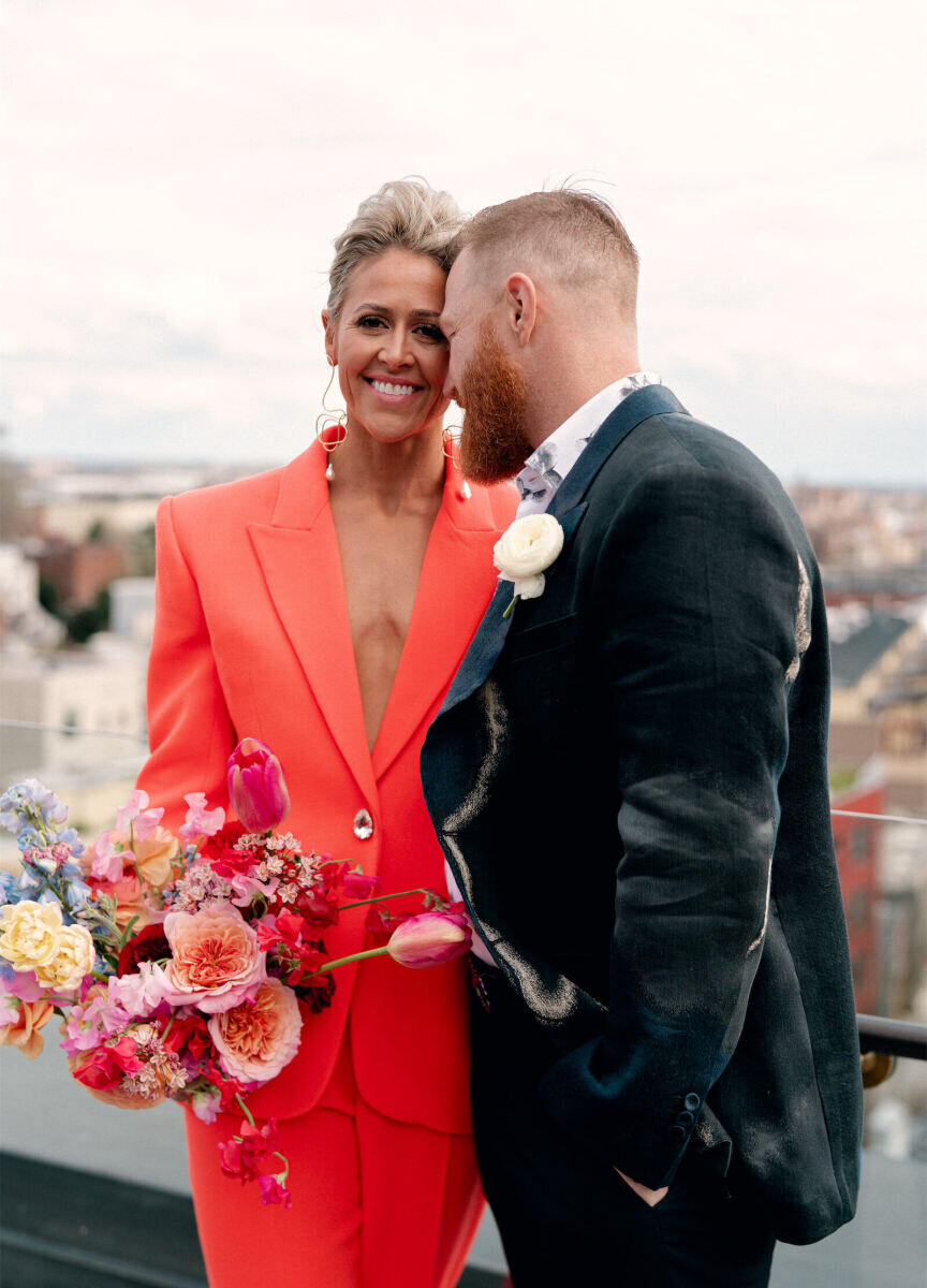 Wedding Dress Shopping: A bride in an orange suit and colorful bouquet on a rooftop with her groom in a suit.