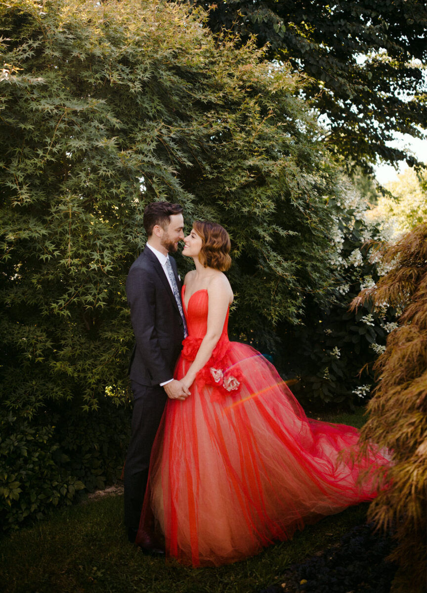 Wedding Dress Shopping: A bride in a red dress holding hands while facing the groom, and they're both surrounded by greenery.