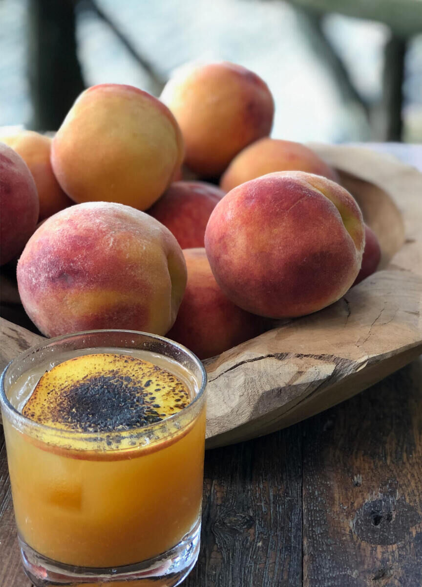 Wedding Drinks: A peach and bourbon cocktail on a wooden table with a wooden bowl full of peaches behind it.