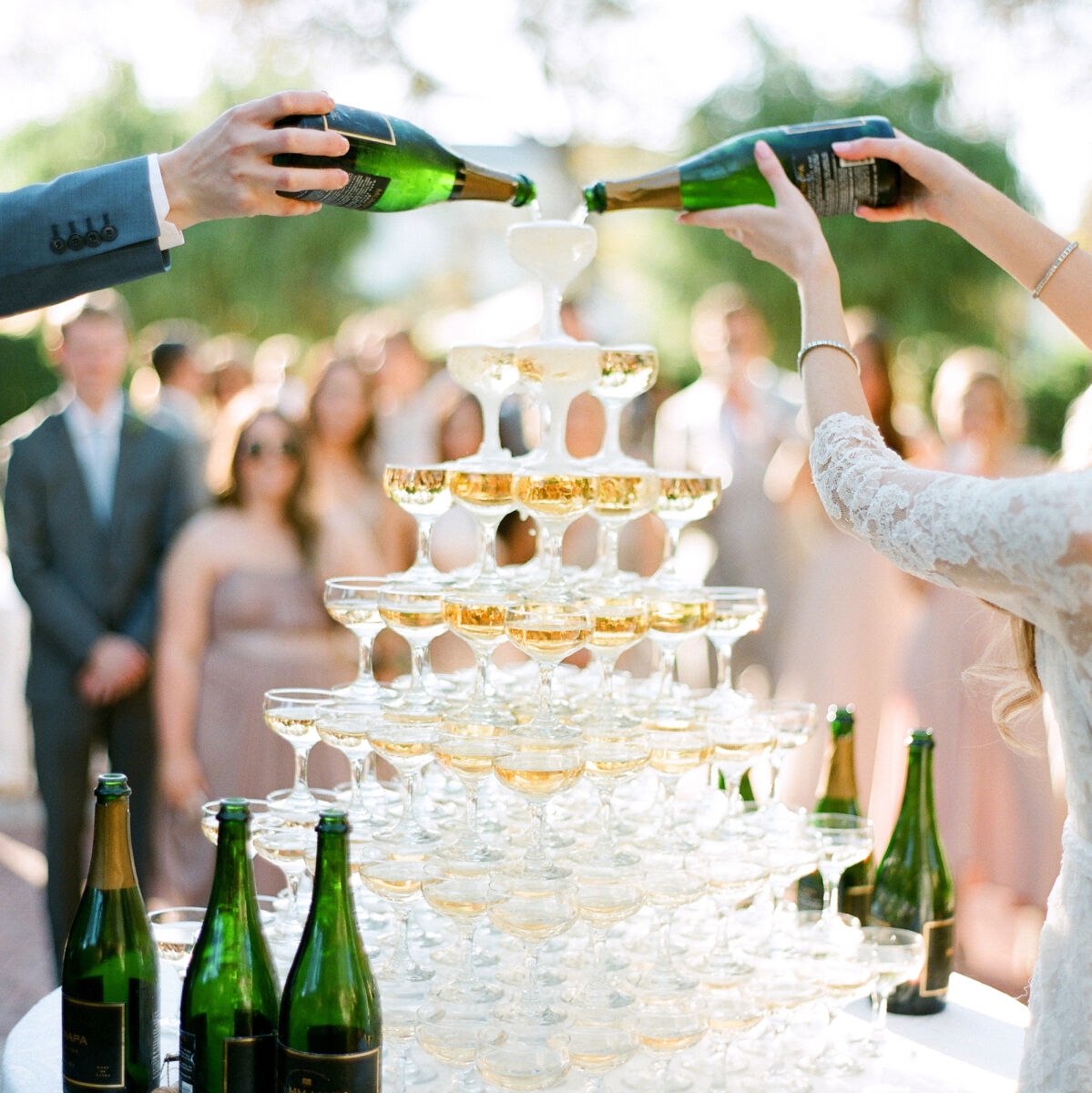 Wedding Etiquette Budget: A groom and bride pouring two bottles of champagne onto a tower of glasses at an outdoor reception.