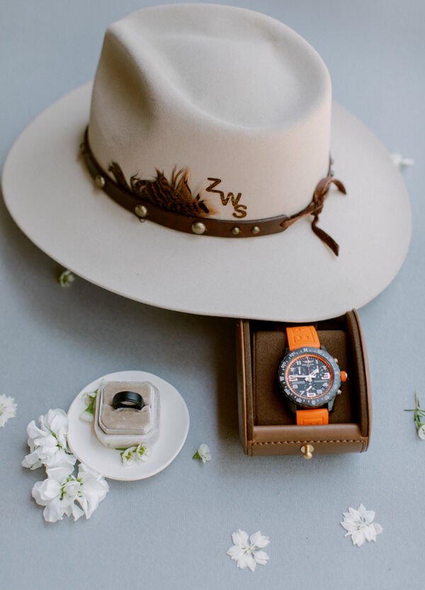 Wedding Etiquette Budget: Monogrammed cowboy hat next to the groom's orange-banded watch and carbon black wedding ring in a cream ring box.