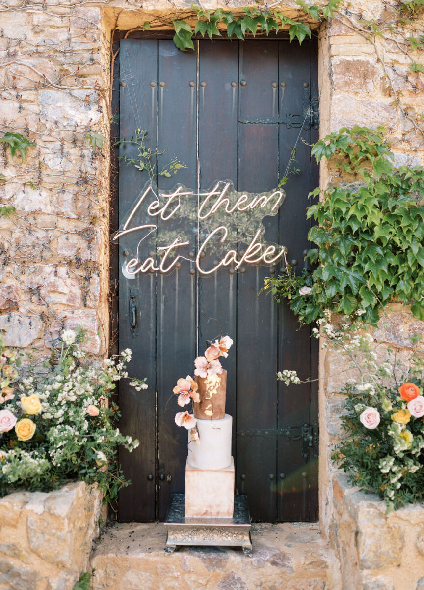 Wedding Etiquette Budget: Gold and cream three-tiered wedding cake in a brick-lined doorway with a black wooden door, adorned with a neon let them eat cake sign.