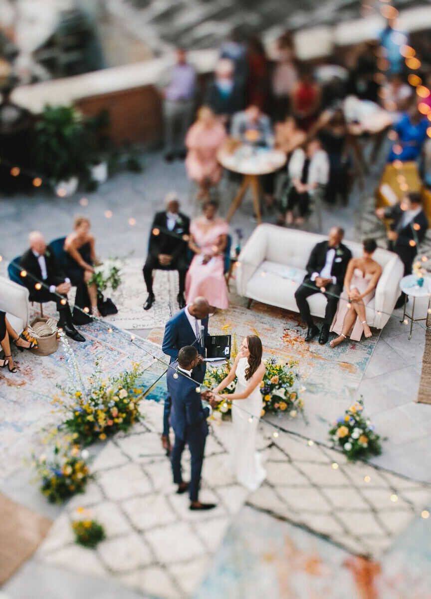 Wedding Photography Ideas: A bird's-eye view of a rooftop, where a wedding couple is standing on a carpet as guests watch from couches, high-top tables, and other areas.