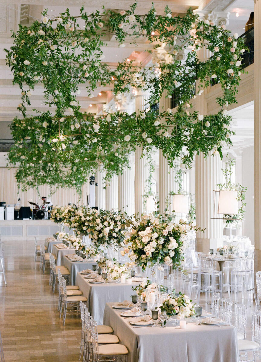 Wedding Photography Ideas: A reception setup with rectangular tables, acrylic chairs, tall white and green centerpieces, and a hanging floral and greenery installation.