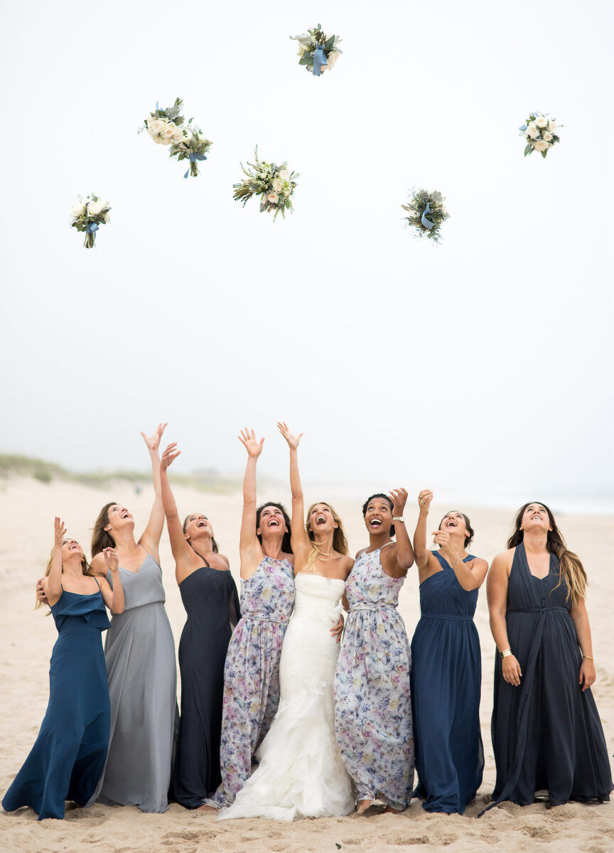 Wedding Photography Ideas: A bride and her bridesmaids, who are wearing various shades of blue, tossing their bouquets high into the air on a beach.