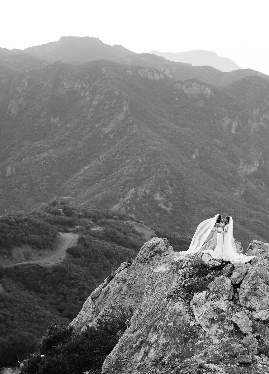 Wedding Photography Ideas: An overhead view of two brides looking at each other on a mountain with a larger mountain range beyond.