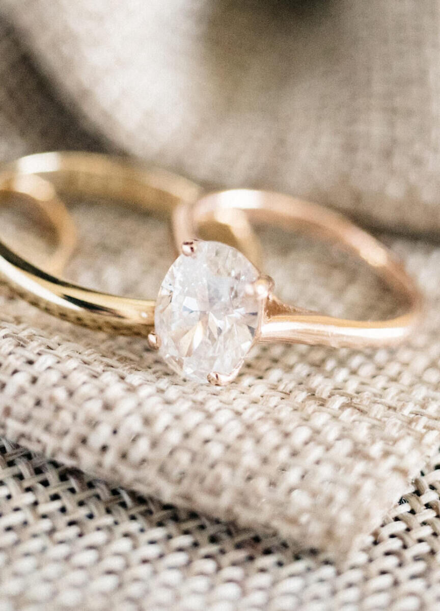 Wedding Ring Design: An oval cut engagement ring in a solitaire sitting perched on beige cloth next to a gold metal wedding band,