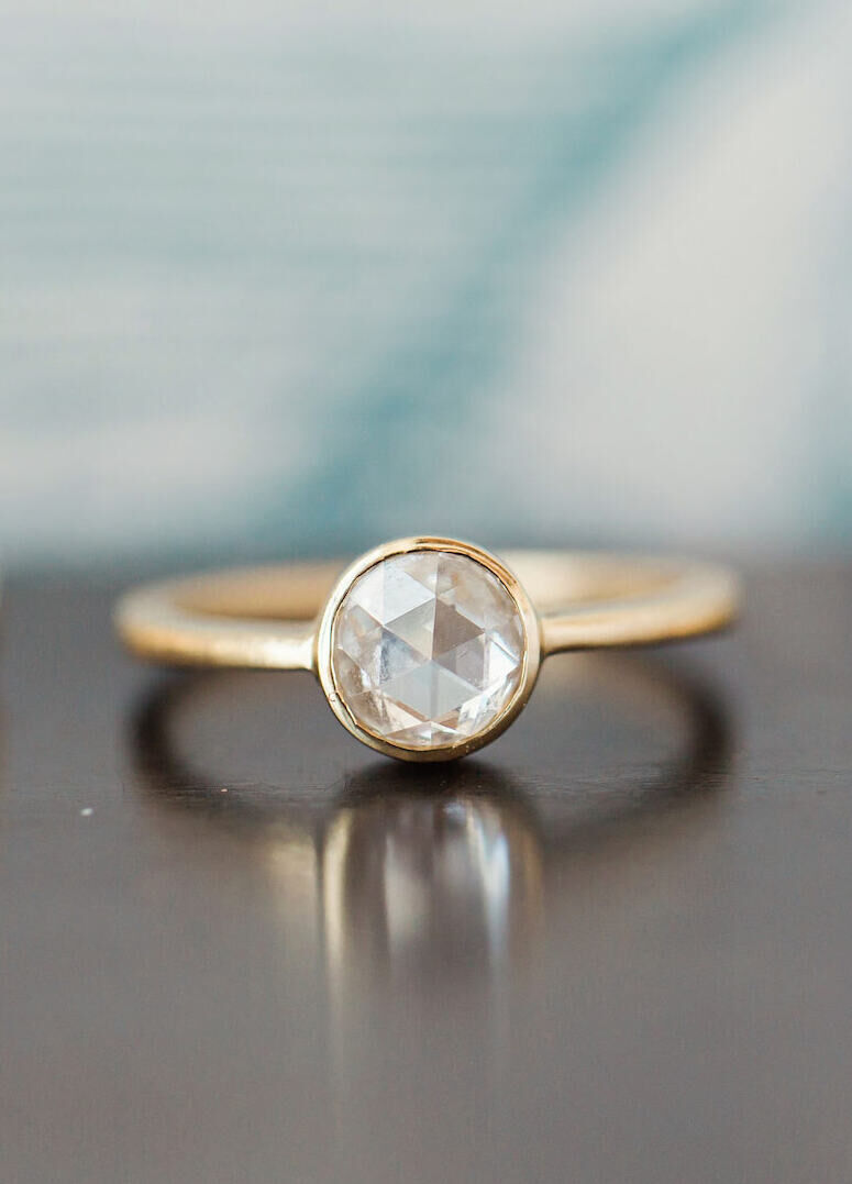 Wedding Ring Design: A round cut diamond in a gold bezel setting resting on a table.