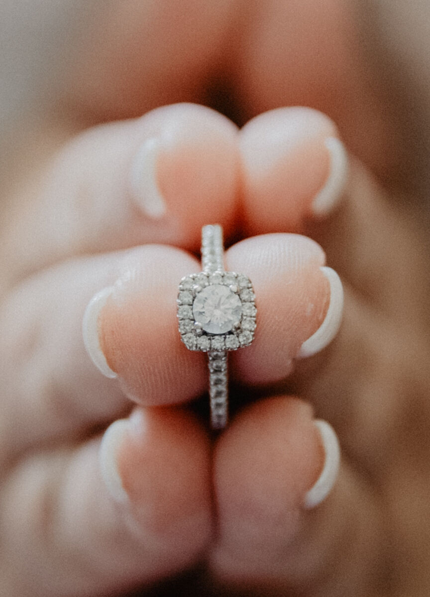 Wedding Ring Design: Two hands holding up a round cut engagement ring with a square halo setting.