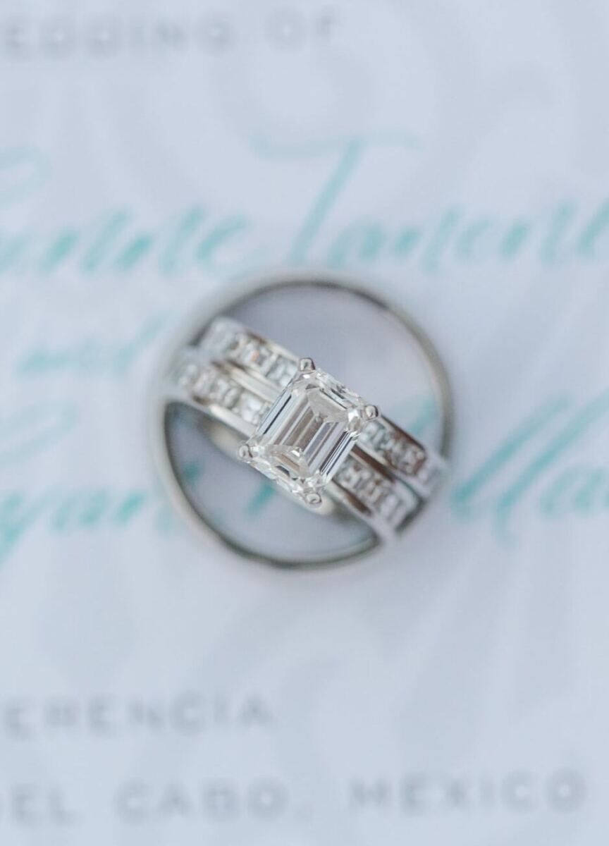 Wedding Ring Design: An emerald cut engagement ring with a channel setting facing up while resting in a wedding band on a blue invitation.