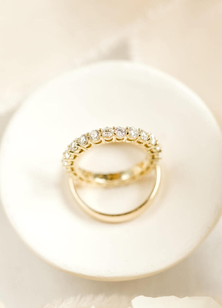 Wedding Ring Design: A gold eternity wedding band sitting in a circular white dish with a gold metal wedding band.