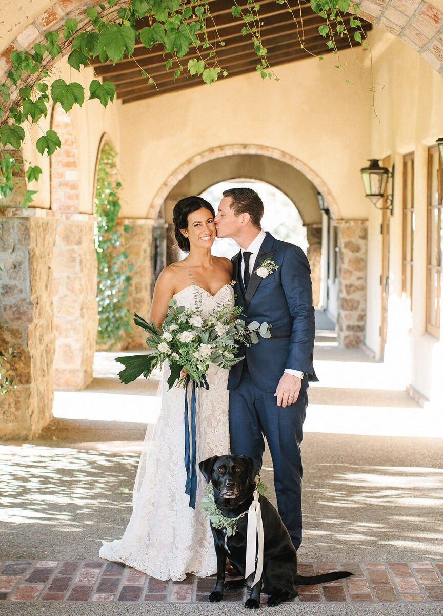 Wedding Venues in California: A groom kissing a bride while posing with their dog in a Spanish-style patio.