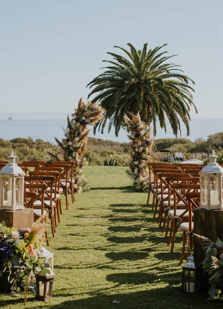 Wedding Venues in California: An outdoor ceremony setup with a palm tree, asymmetrical arch, and wooden chairs overlooking the ocean.