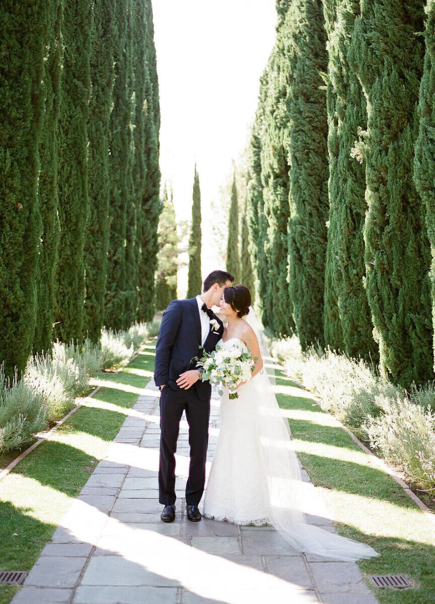 Wedding Venues in California: A groom kissing a bride along a pathway lined with trees.