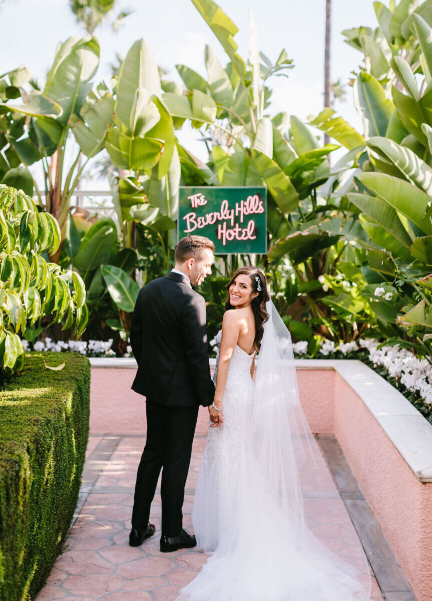 Wedding Venues in California: A bride looking toward the camera while a groom looks at her in a palm-covered area with a Beverly Hills Hotel sign in the background.