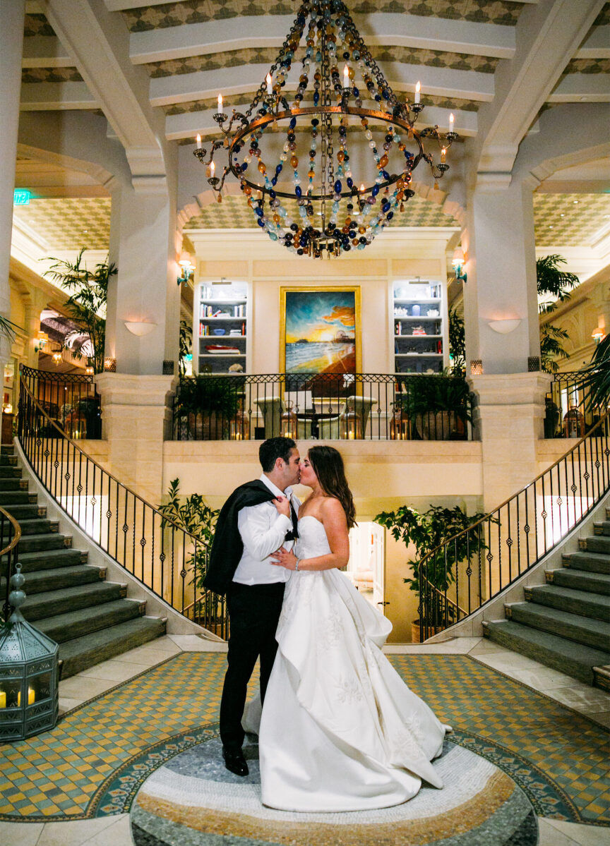 Wedding Venues in California: A wedding couple kissing at the bottom of a staircase with an oversized chandelier above.