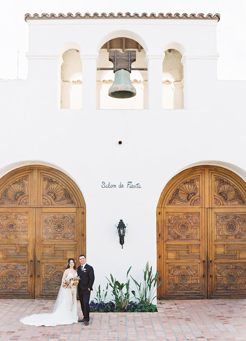 Wedding Venues in California: A wedding couple posing in front of a Spanish-style building with a bell and large wooden doors.