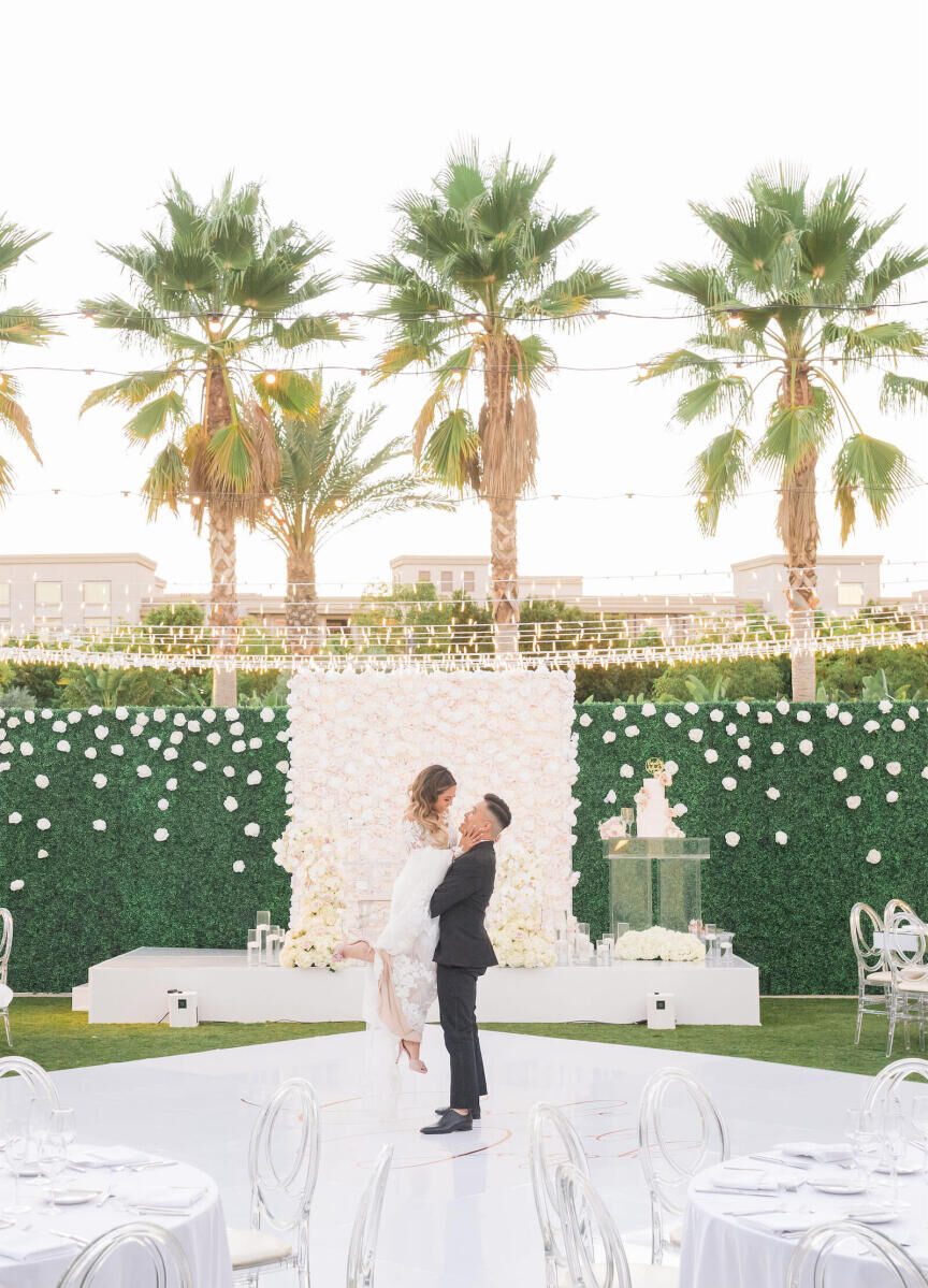 Wedding Venues in California: A groom picking up a bride at an outdoor reception area with string lights above them and palm trees in the background.