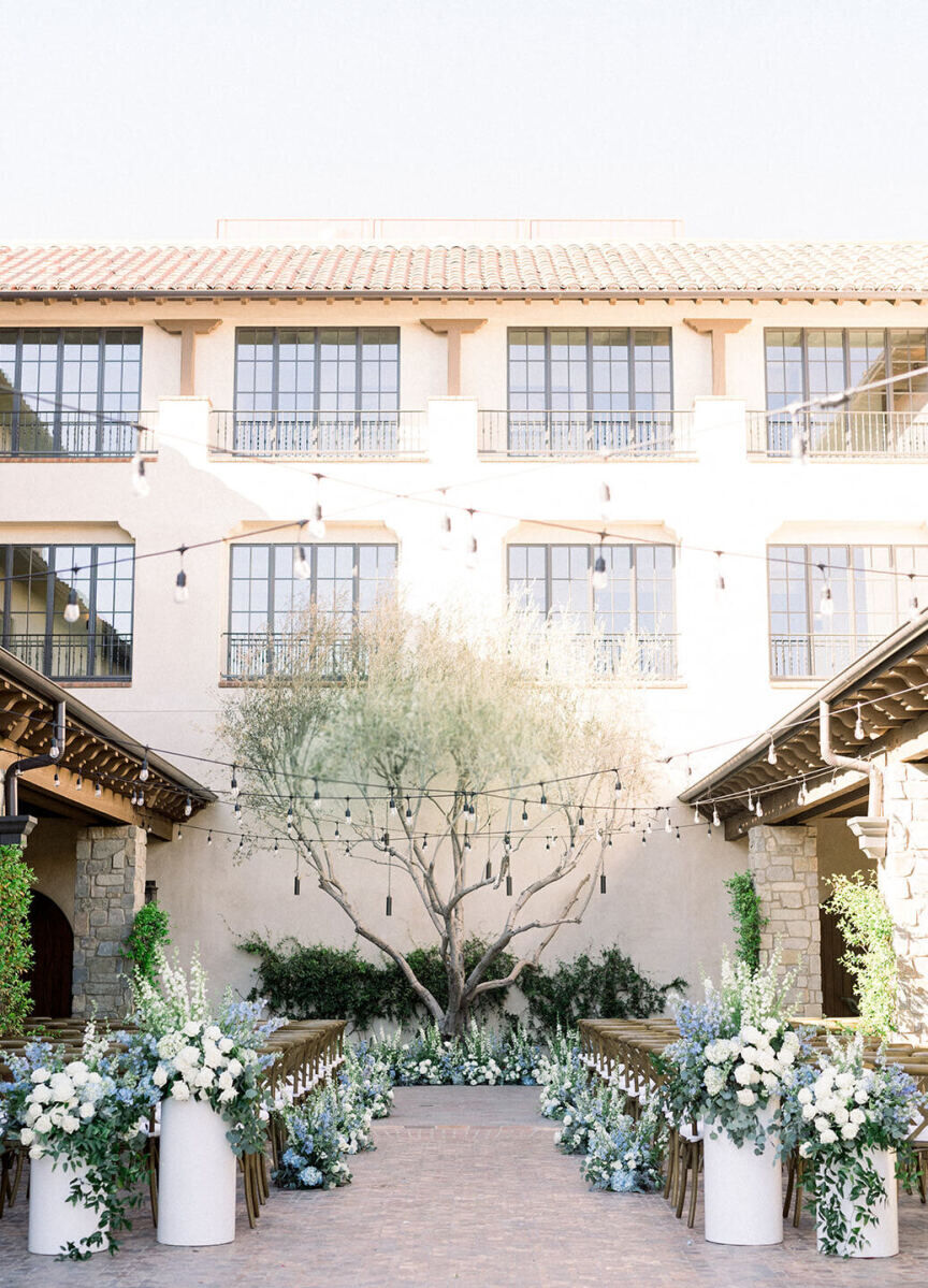 Wedding Venues in California: An outdoor ceremony setup with white and blue floral arrangements leading down the aisle to a tree in a Spanish-style courtyard.