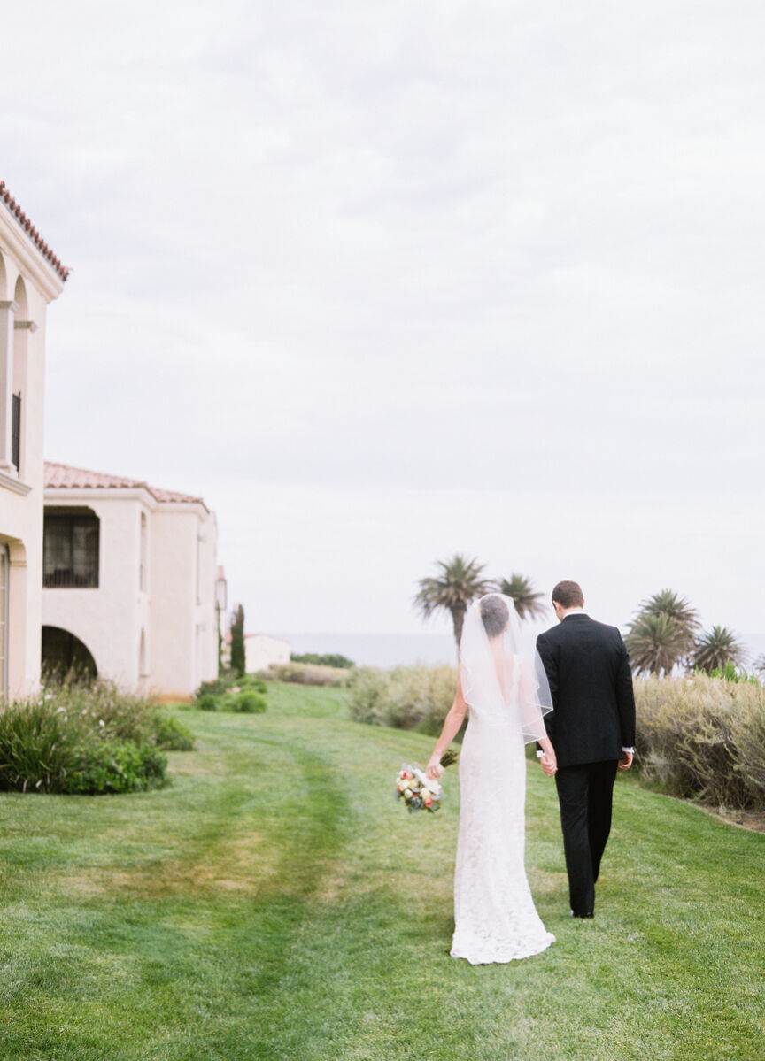 Wedding Venues in California: A wedding couple walking on a grassy area with a Spanish-style building to the left and the ocean to the right.