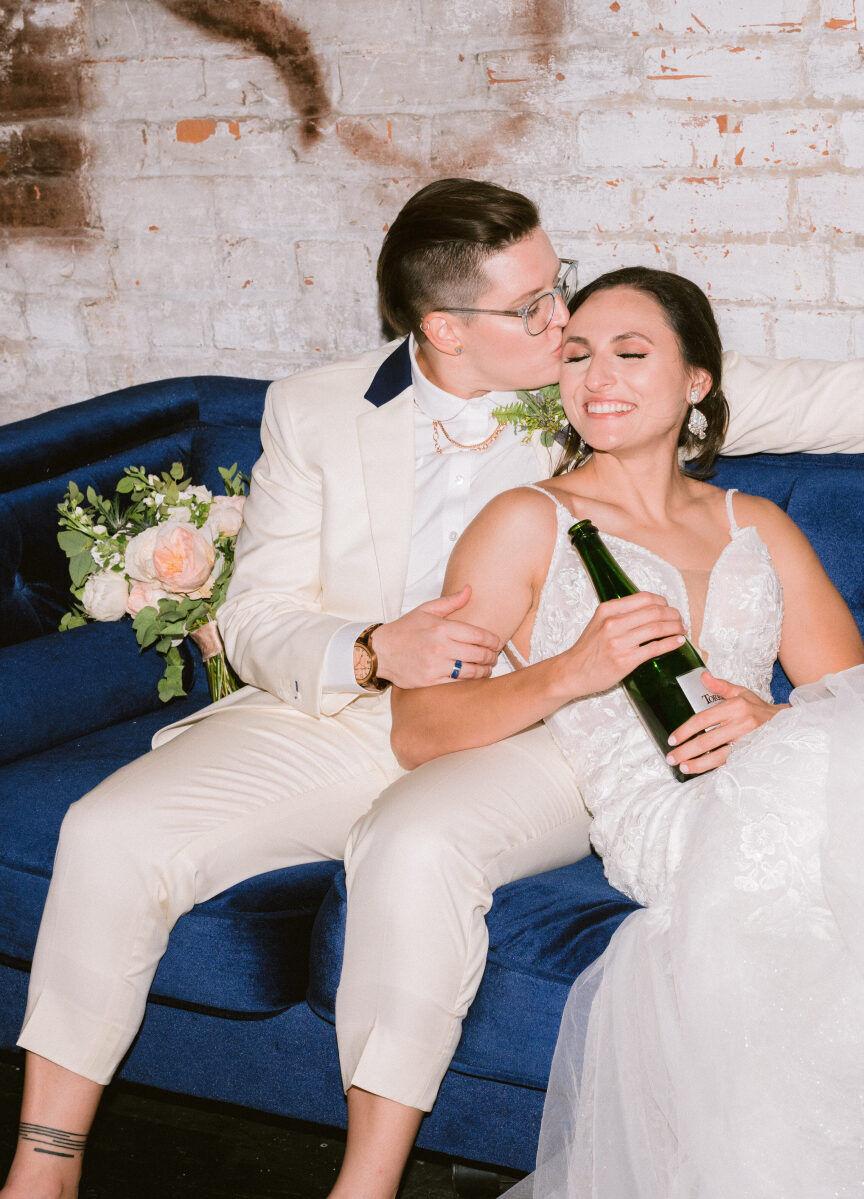 Wedding Website Examples: A wedding couple sharing a moment on a navy couch with a bouquet and bottle of wine.