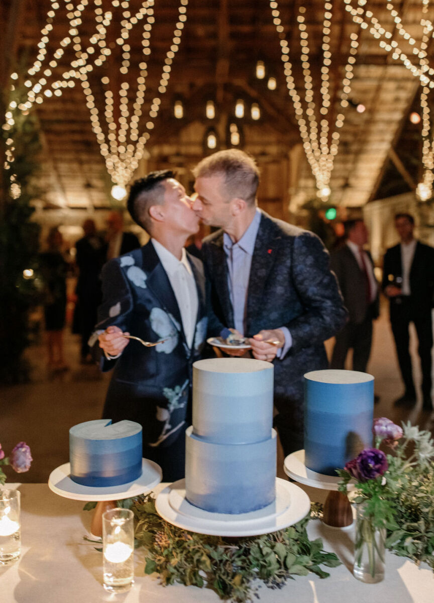 Wedding Website Examples: A wedding couple sharing a kiss by their three blue wedding cakes at an indoor reception.