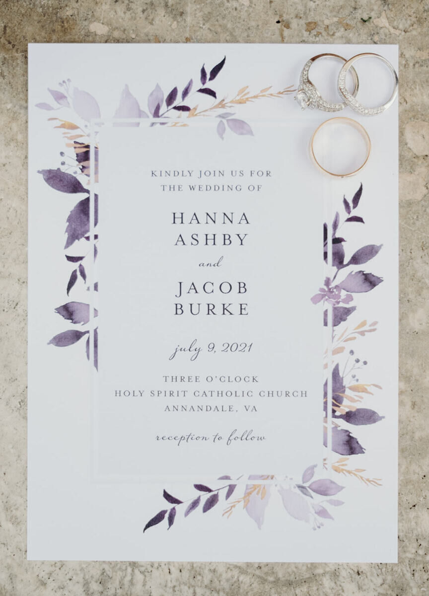 Wedding Website Examples: A flat lay showing a lilac-accented white wedding invitation with an engagement ring and wedding rings.