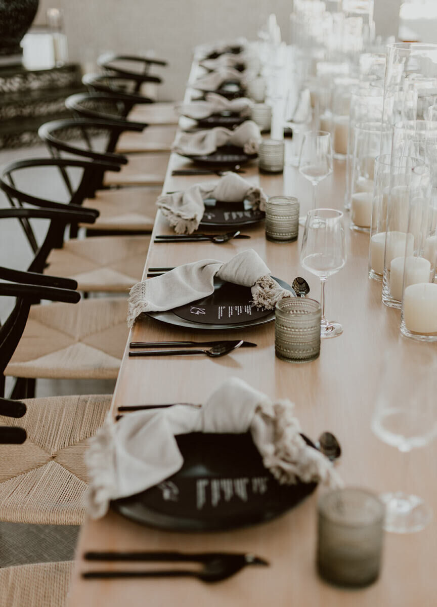 A modern, Cabo wedding reception, with a long wood table set with black chairs, knotted white napkins, and black flatware.