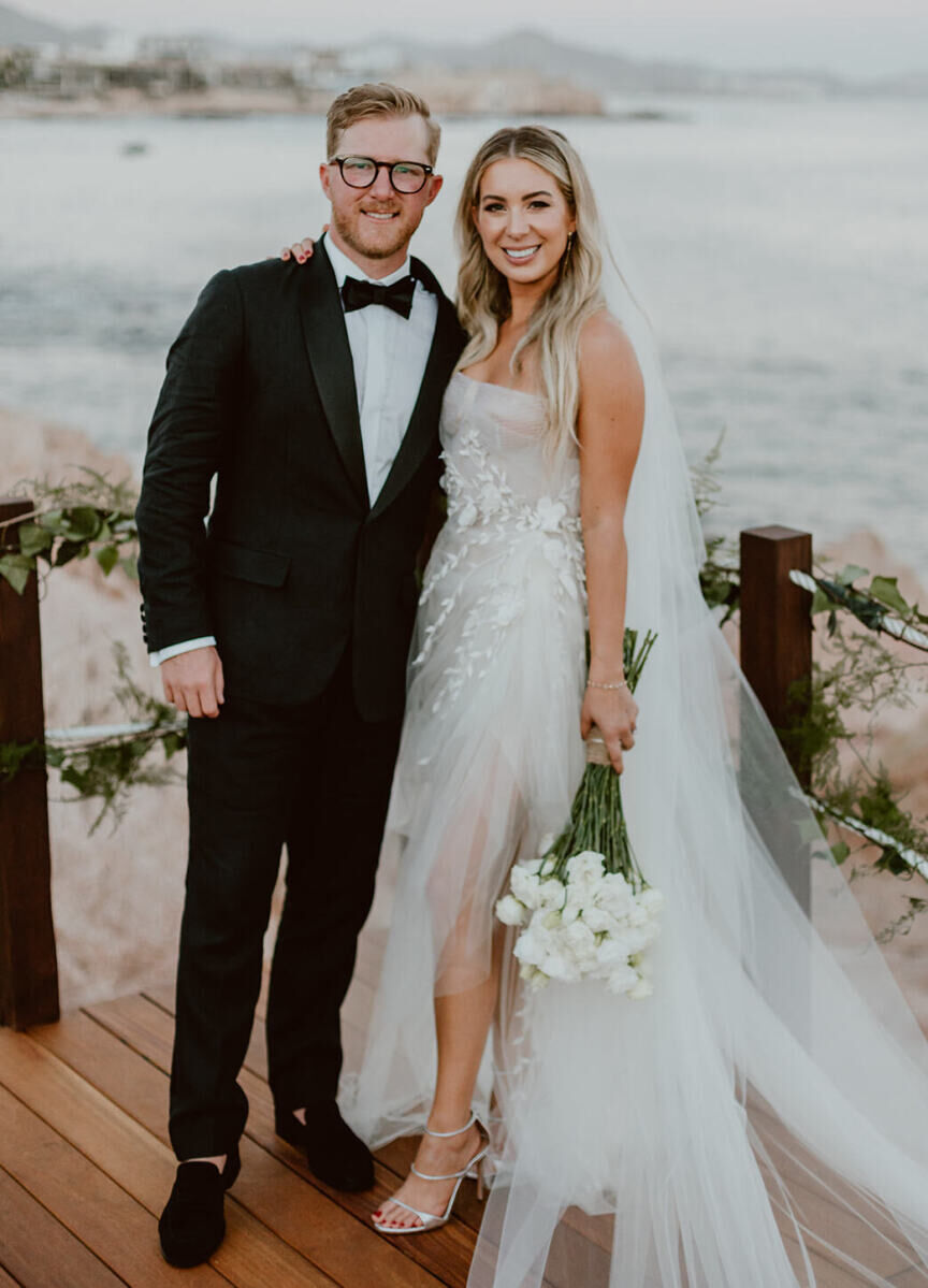 A groom in a tuxedo and a bride in a high-low wedding dress, holding her bouquet of white roses while standing on a platform near the beach of Cabo.