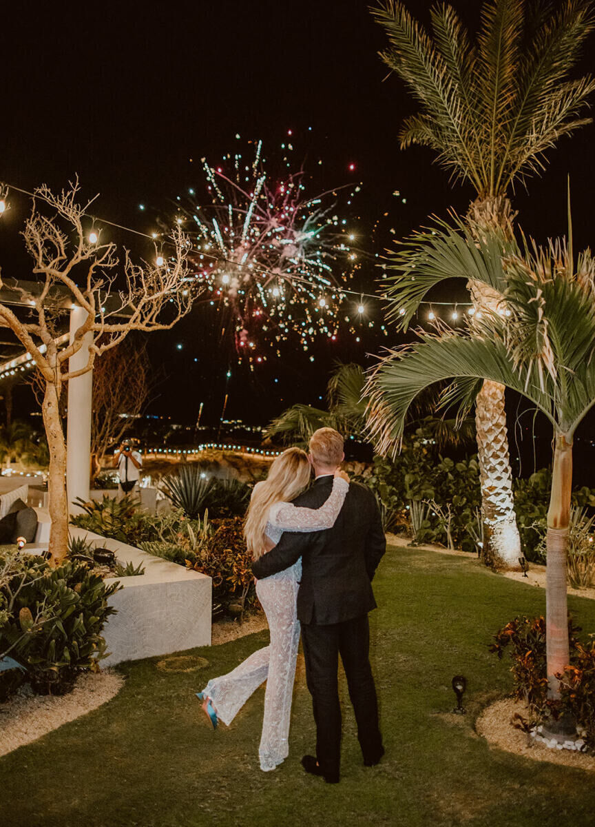 A bride in her reception jumpsuit embraces her groom, while taking in a fireworks display during their Cabo wedding.