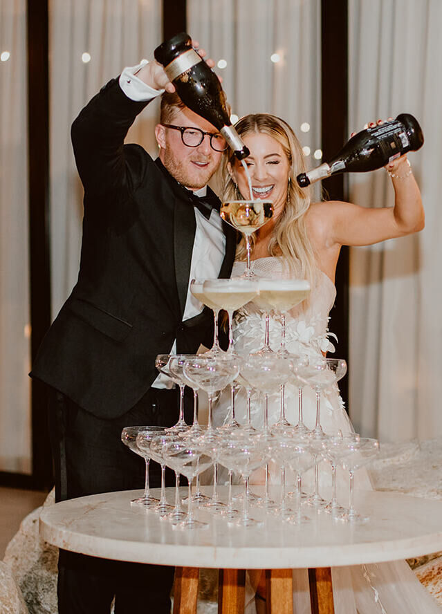 A bride and groom pour Champagne over a tower of coupe glasses.