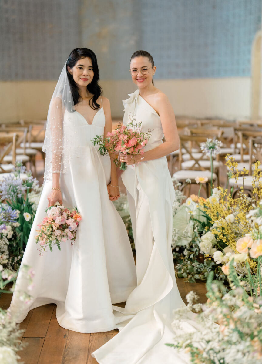 A pair of brides holding pink bouquets smile at the setting of their whimsical wedding ceremony, surrounded by a curved installation of pastel flowers.