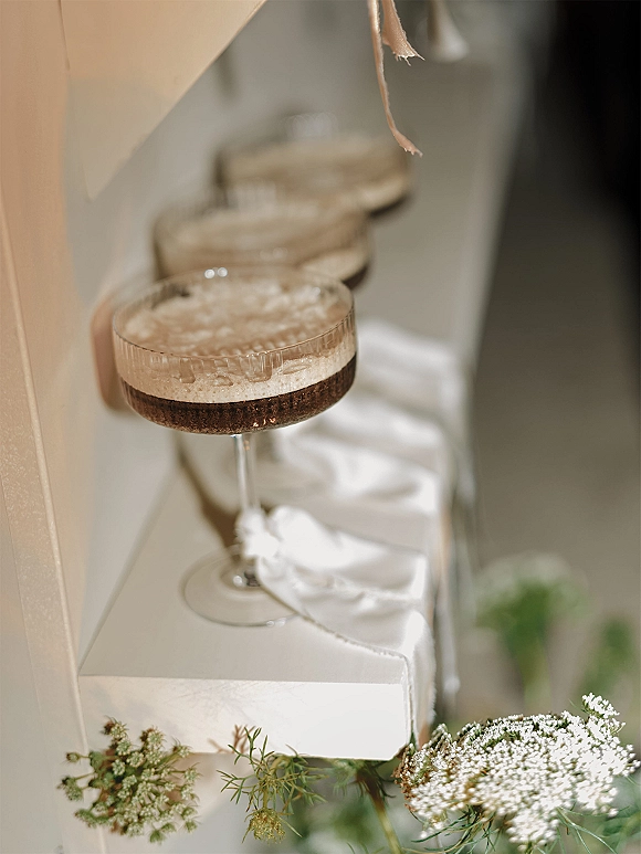 Wedding cocktails in champagne coupe glasses with foamy tops and baby’s breath accents, lined on a ledge against a neutral wall in soft light