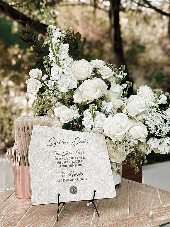 Signature drinks sign with wedding signature drinks listed beside white roses and greenery on a wood bar top with copper cups outdoors