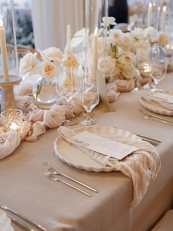 Reception tablescape with wedding place setting, scalloped plates, menu card, white rose centerpieces, and ivory taper candles in clear holders indoors