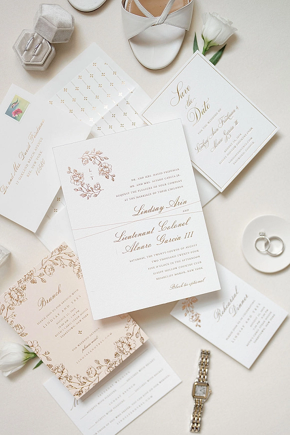 Invitation suite wedding invitation flat lay with gold foil calligraphy, wax seal envelope, rings, perfume, watch, and white rose buds on neutral tabletop