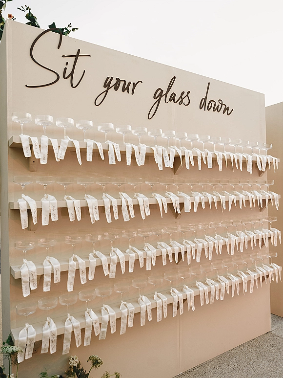 Wedding seating chart escort card wall with champagne coupe glasses, ribbon tags, and calligraphy lettering on shelves against a neutral wall
