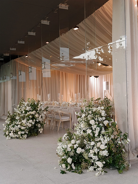 Reception decor with white floral arrangements and greenery lining long banquet tables, candles glowing under a draped fabric ceiling in a modern venue