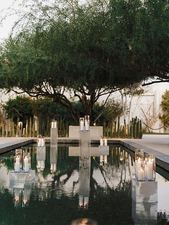 Pool wedding decor with floating candle centerpieces in glass cylinders on concrete pedestals lining a reflecting pool in a courtyard with cacti