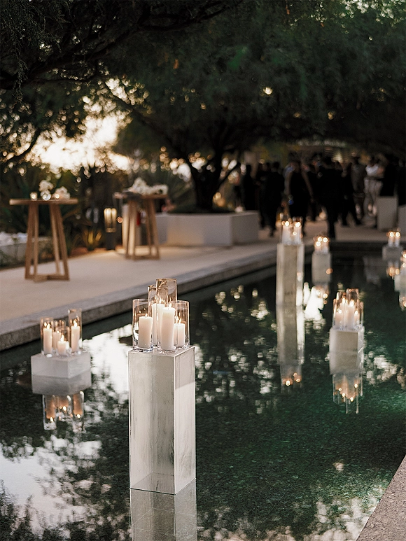 Poolside wedding decor with floating candle centerpieces in glass hurricane vases and white pillar candles glowing beside a reflecting pool on a terrace